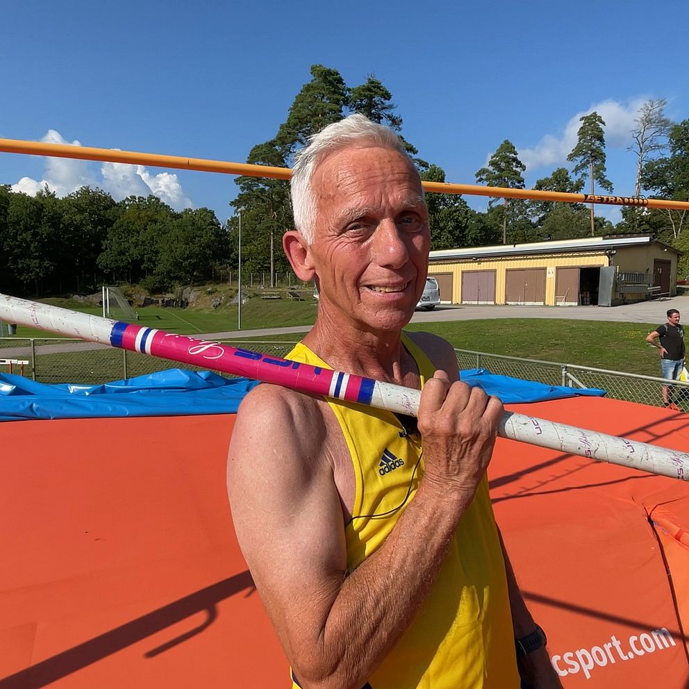 En man i idrottskläder står på en idrottsplats med en stavhopps-stav över axeln