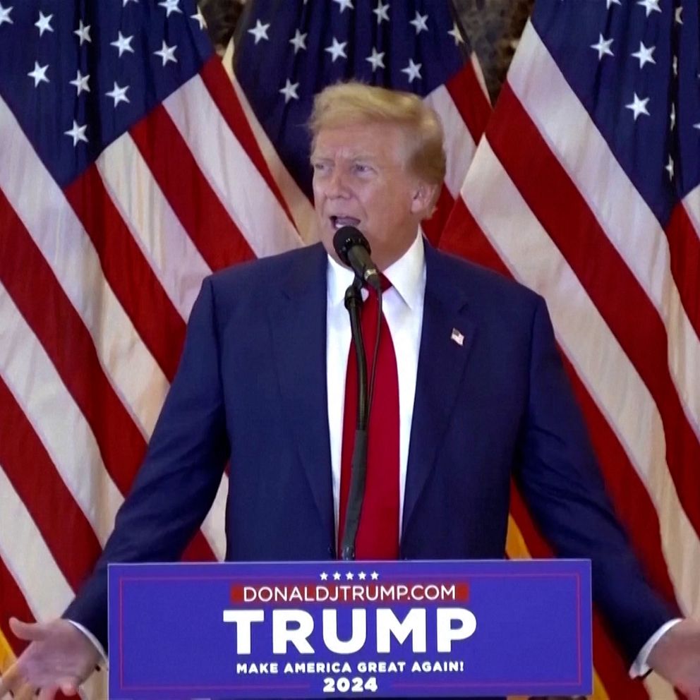 En man står vid ett podium med en skylt som har texten ”TRUMP 2024” och ”MAKE AMERICA GREAT AGAIN”, omgiven av amerikanska flaggor. Han har klädd i en mörk kostym med en röd slips och gestikulerar med händerna.