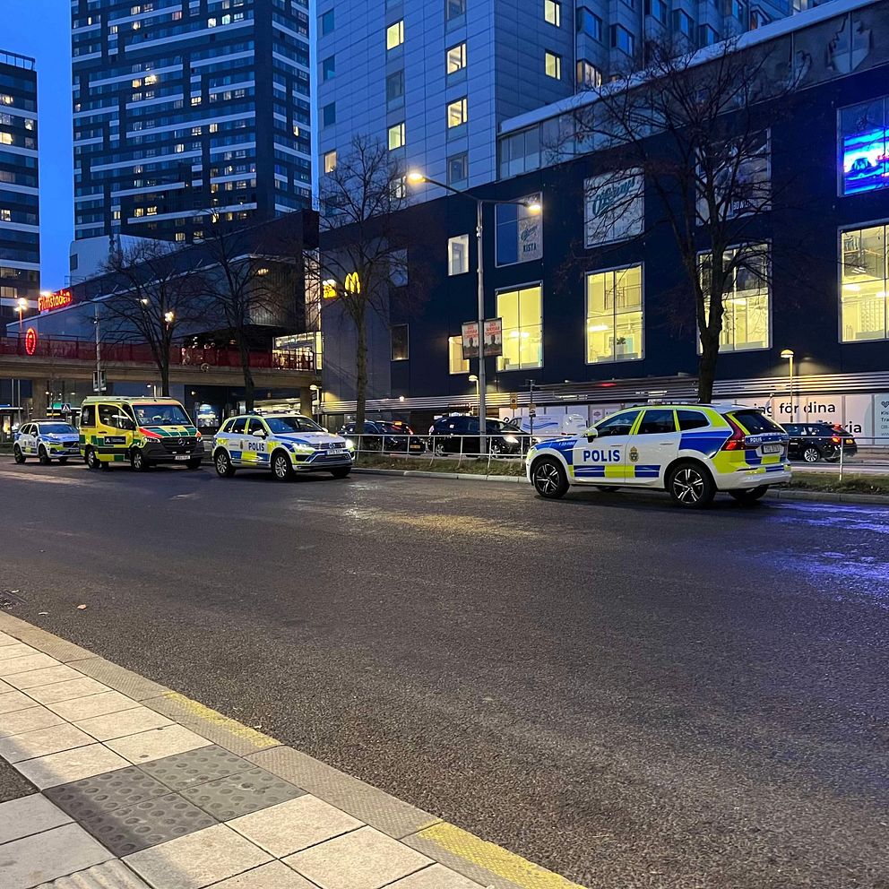 Polisfordon står parkerade längs en gata i en stadsmiljö under skymning. I bakgrunden syns höga byggnader och en McDonald's-restaurang. Belysningen från byggnaderna och gatlyktorna ger en dämpad atmosfär.