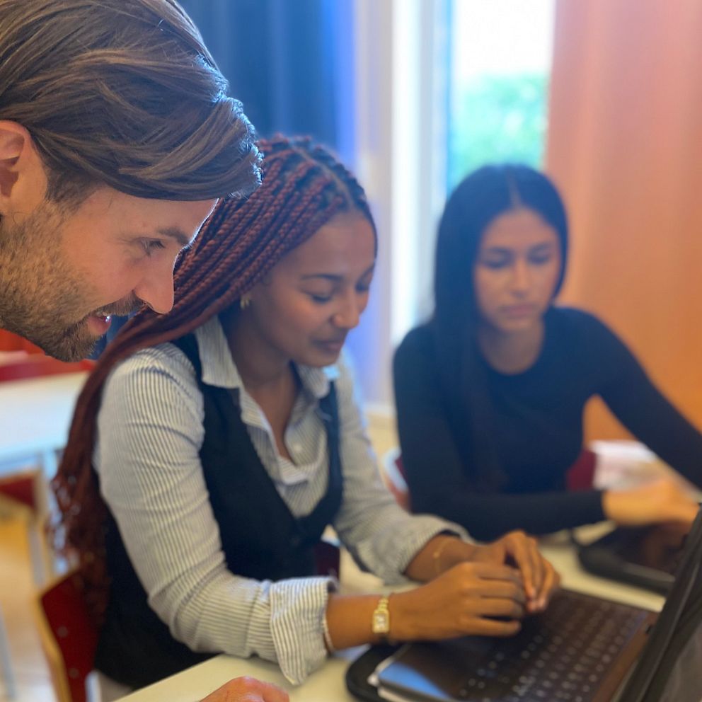 Tre personer vid ett bord med bärbara datorer. Läraren Anders Österholm lutar sig framåt. Dina Tebebu Tifera och Alicia Tuguay sitter ner och ser på skärmen. Rummet har naturligt ljus som strömmar in genom ett fönster, och det finns stolar och bord i bakgrunden. Atmosfären verkar fokuserad och samarbetande.