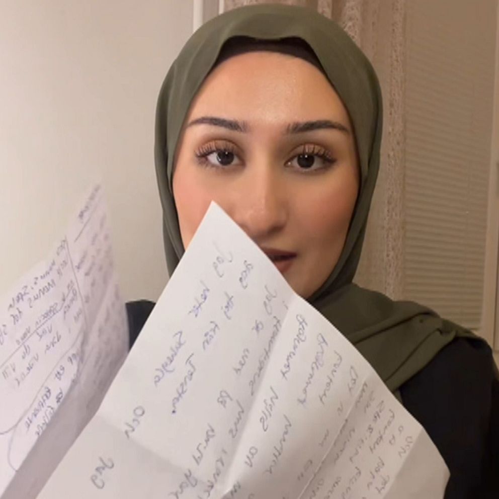 En person som håller upp flera handskrivna papper med text, iklädd en grön huvudduk och med en neutral bakgrund.