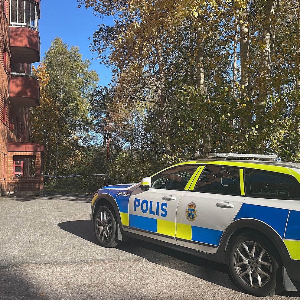 En polisbil står parkerad framför en flerfamiljshus, omgiven av träd med höstliga löv. Byggnaden har röda fönsterkarmar och balkonger.