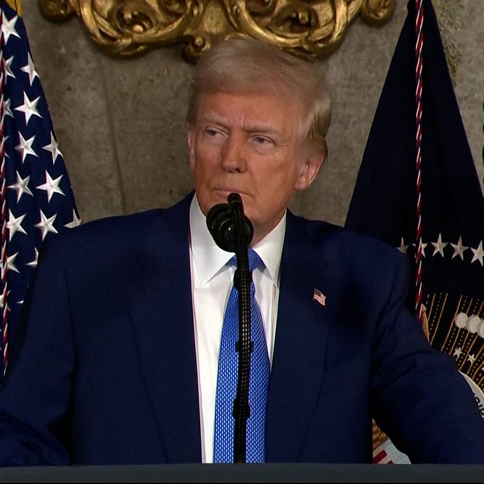Person i kostym talar vid podium, amerikanska flaggor i bakgrunden.
