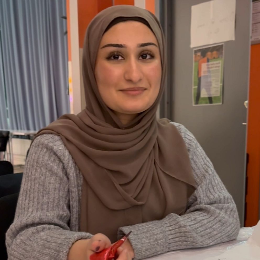 En person med en sjal sittande vid ett bord, håller i en röd föremål. Bakgrunden visar en inomhusmiljö med blåa gardiner och en vägg med affischer.