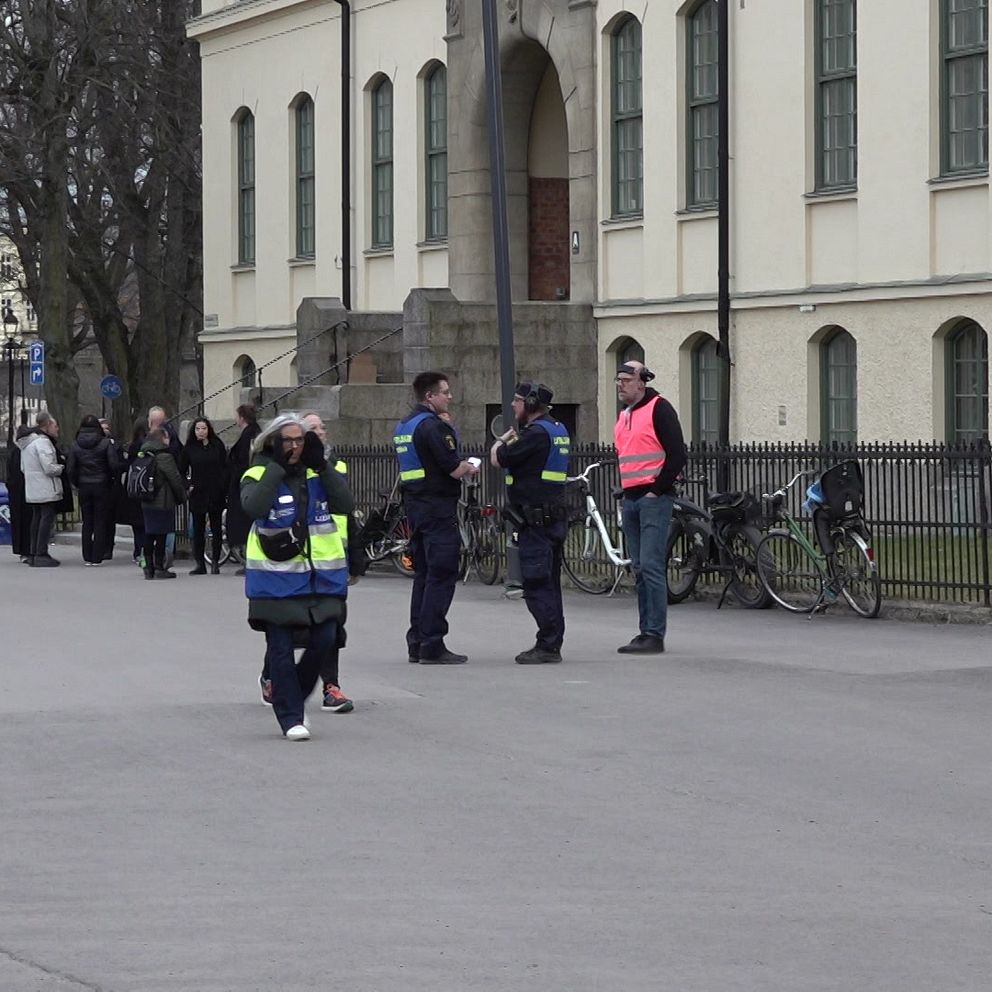 Säkerhetspersonal samlas nära en byggnad. En buss och några cyklar syns i bakgrunden.