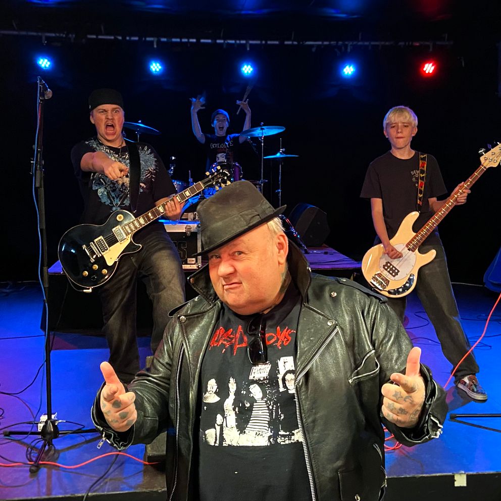 Ett rockband på scen, en person står i förgrunden med tumme upp. Scenen är belyst med färgglada ljus.