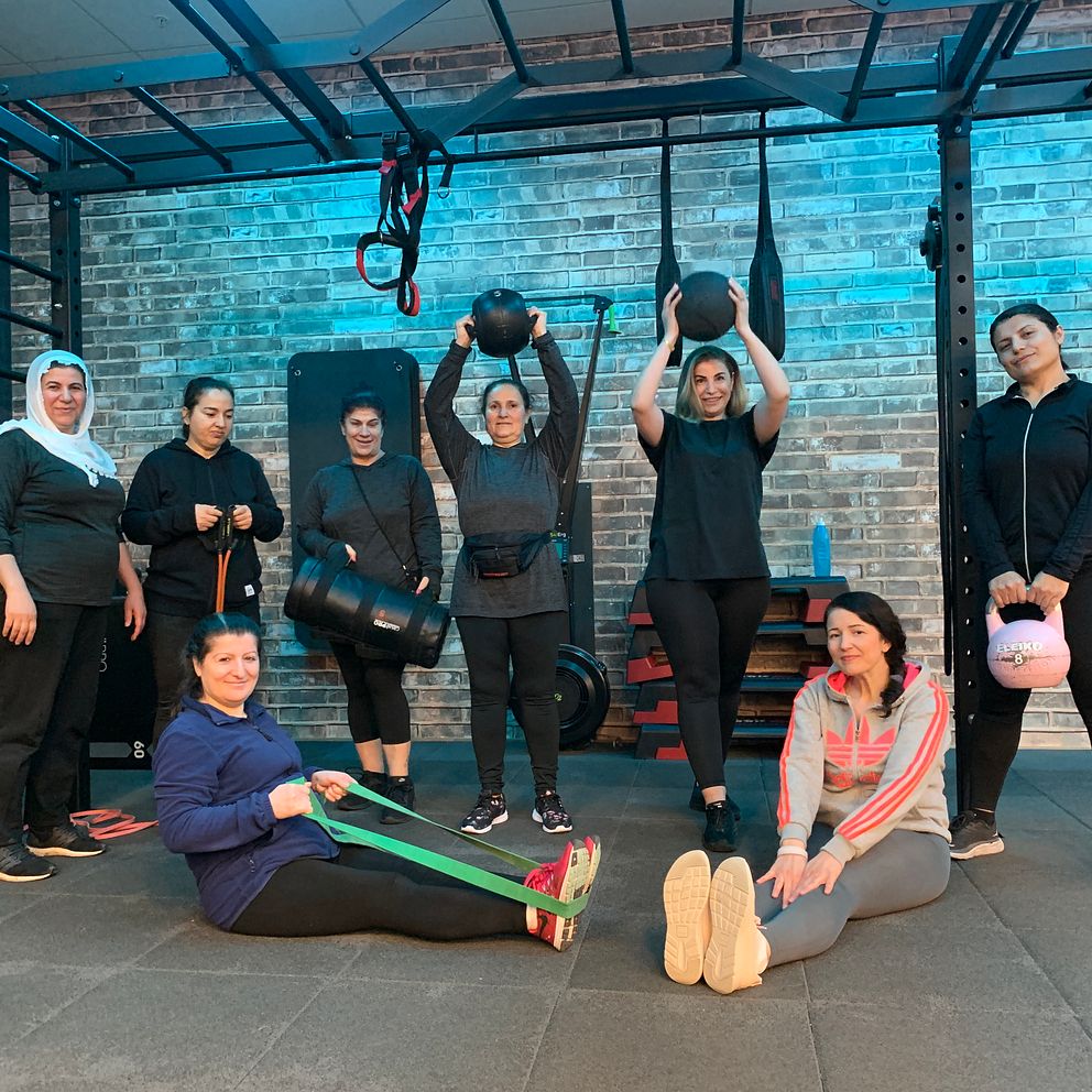 Åtta kvinnor står och sitter på mjuk matta i gym, de håller träningsbollar- och band.