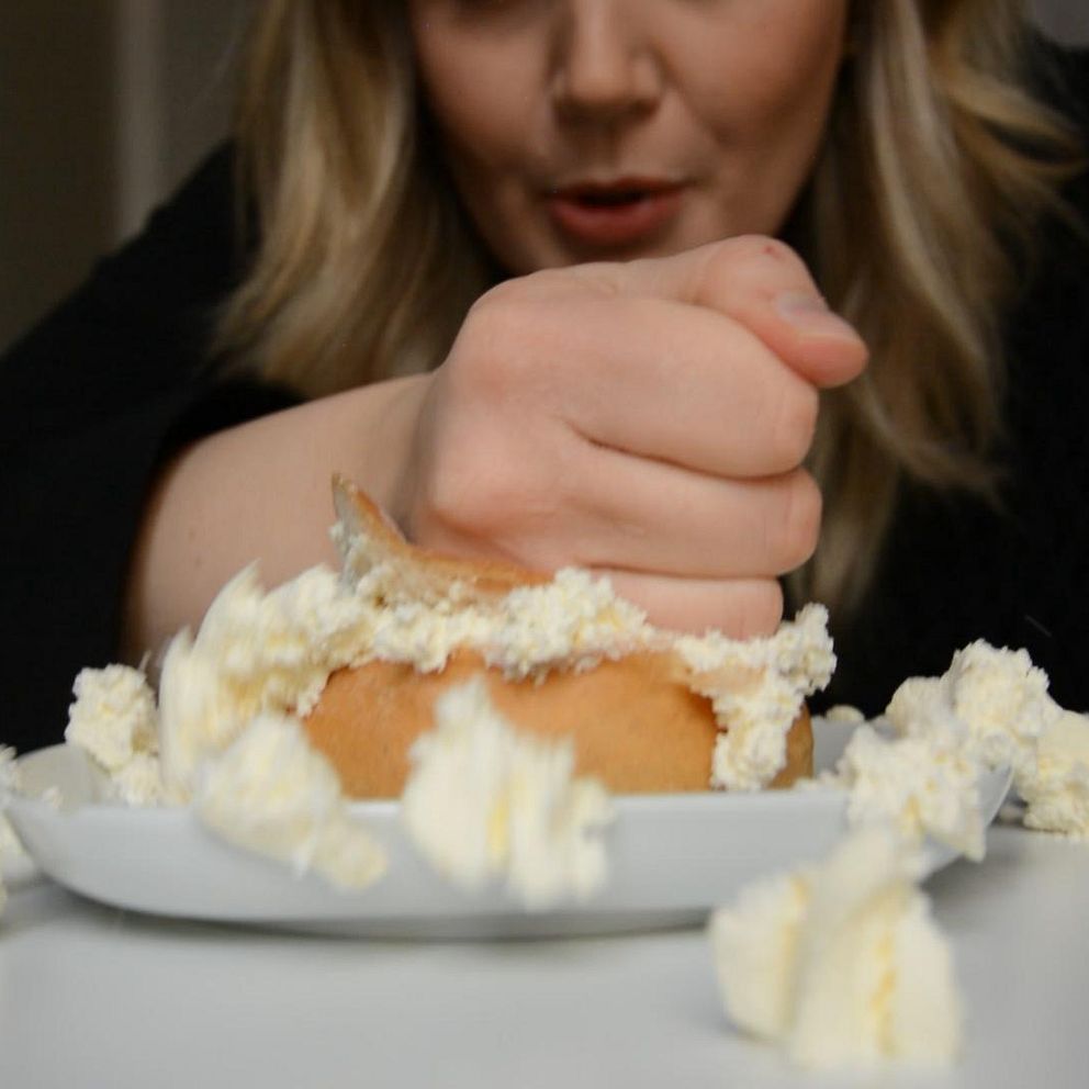 Person trycker ner en hand i en semla, vilket får grädden att spilla ut över tallriken.