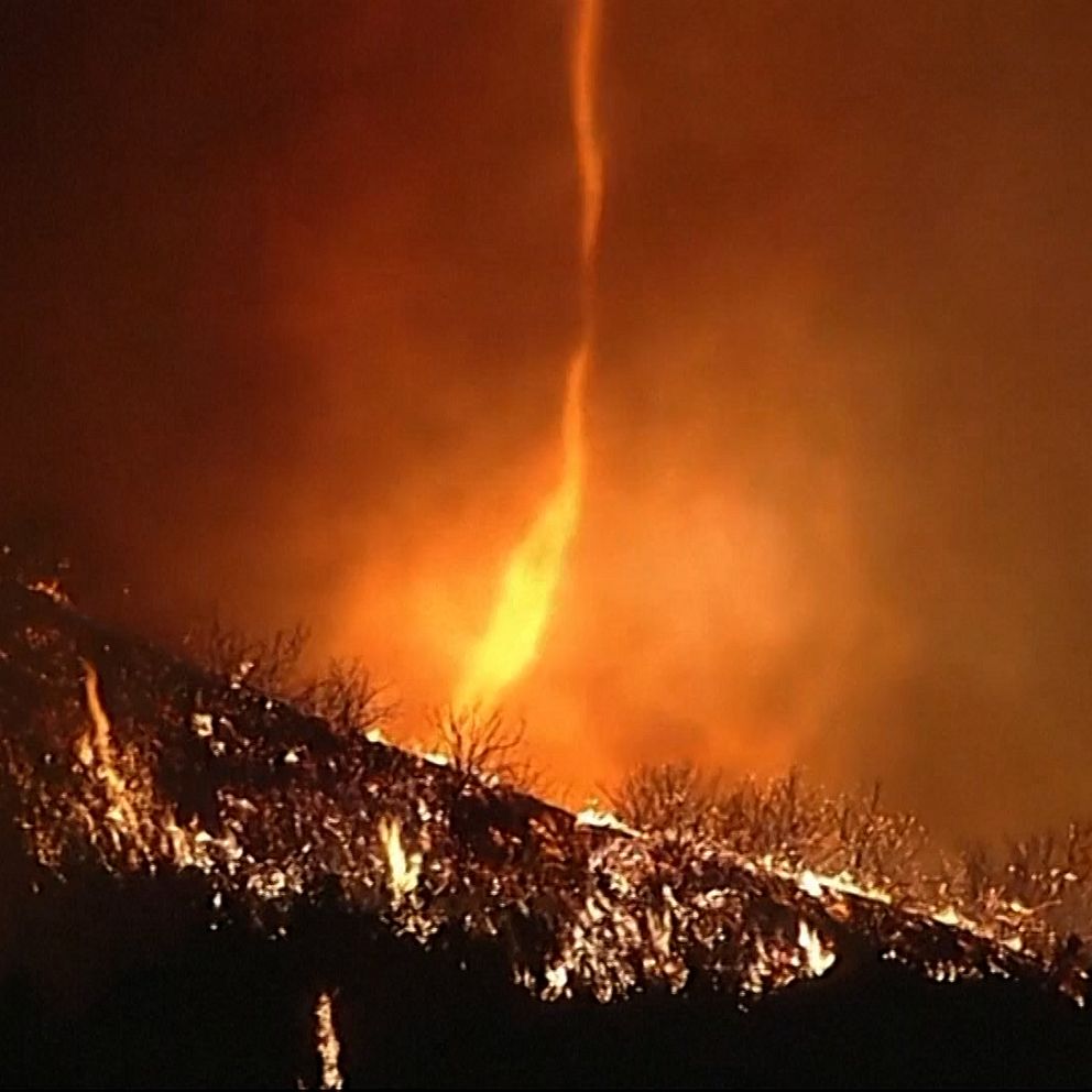 En kraftig eldsvåda tillsammans med en eldtornado rasar över ett bergslandskap, med lågor som slår upp mot himlen och en eldvirvel som transporterar rök och gnistor.
