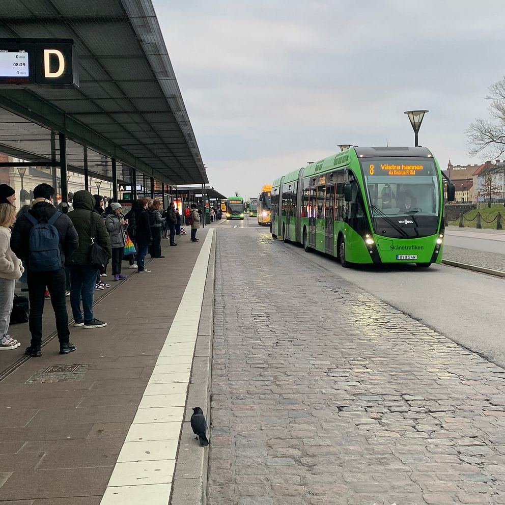 Människor på Malmö C står vid en busshållplats under tak och väntar på en grön, modern stadsbuss som närmar sig i en stadsmiljö med molnigt väder.