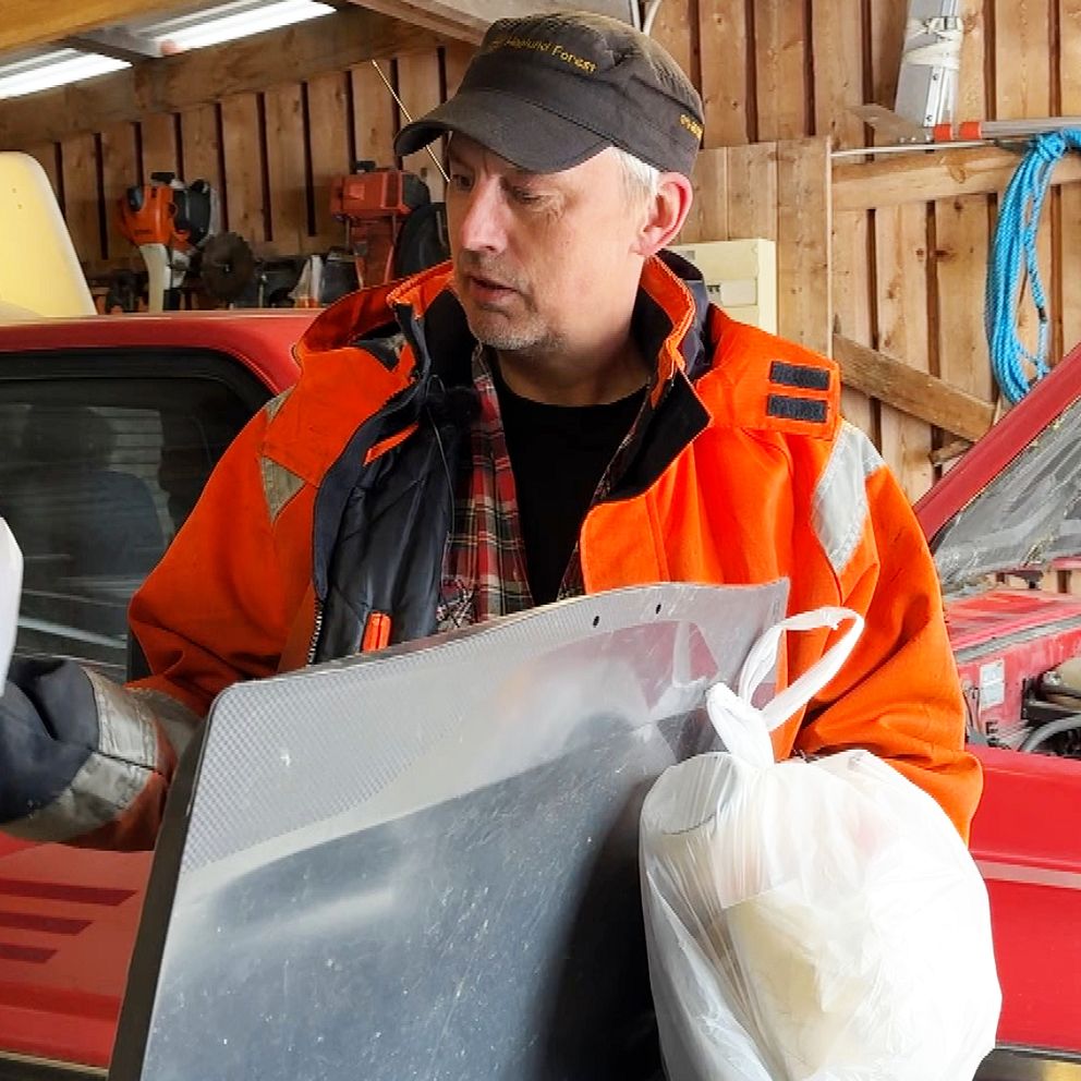 Person i orange jacka håller plastpåsar och bildel framför röd bil med öppen motorhuv.