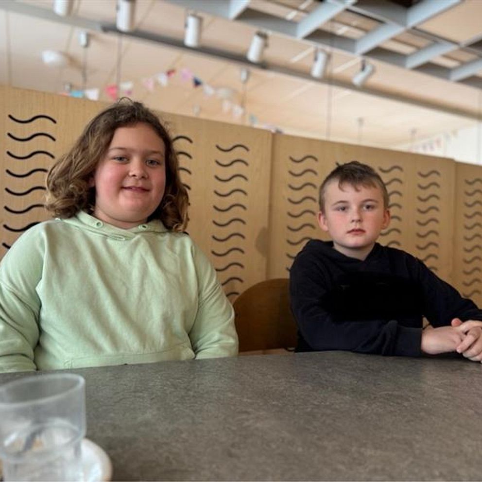 Två personer sitter vid ett bord i en matsal eller kafé, med fokus på deras överkroppar och omgivningen i bakgrunden. På bordet finns ett tomt glas och en tallrik med matrester. Bakom dem ses en dekorativ träskärm och takbelysning.