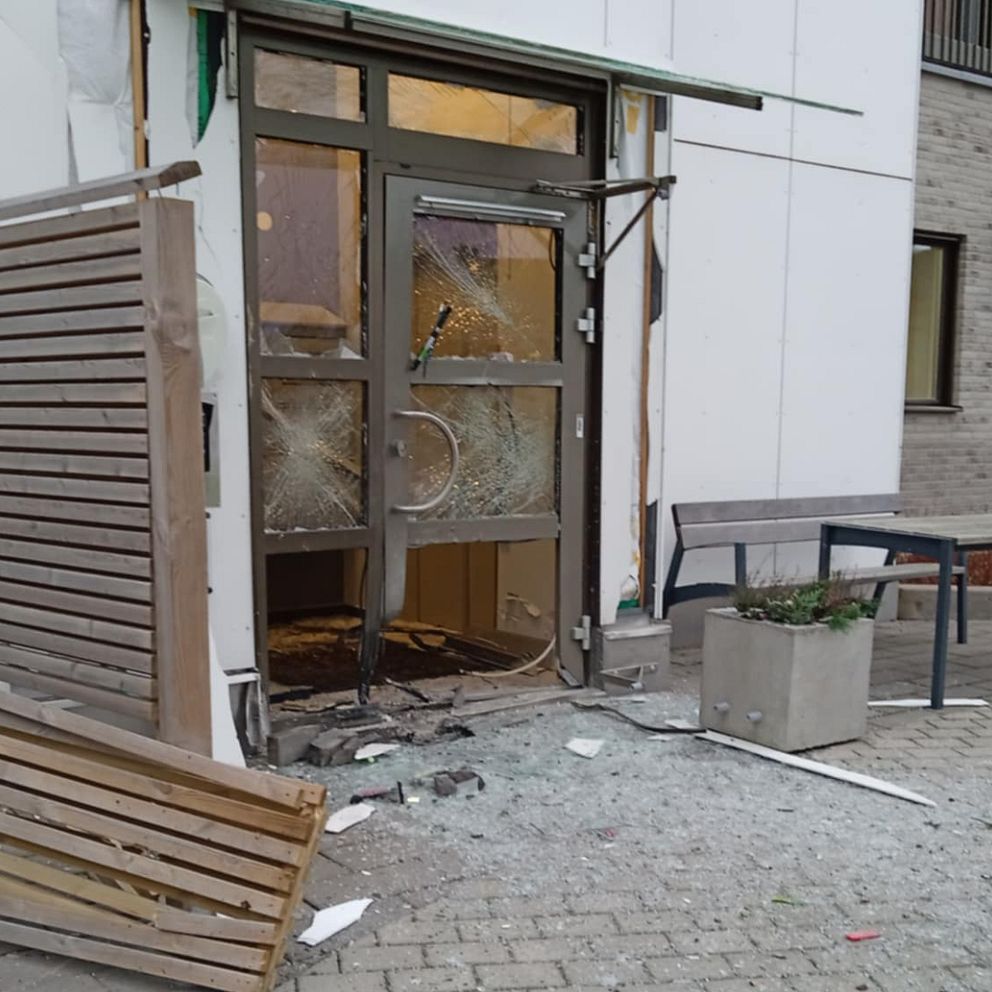 Skadad entré med spruckna fönster och en trasig dörr, där en del av fasaden har lossnat. Det ligger glas och skräp på marken, och en liggande träpanel är synlig vid sidan av dörren. I bakgrunden skymtar en grön växt i en kruka och byggnadens väggar.