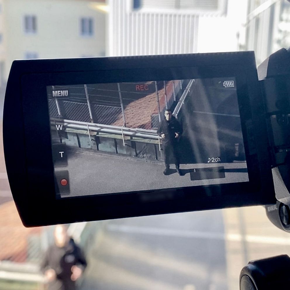 En videokamera är riktad genom ett fönster mot en reporter längre ned på marken, med en kamera-skärm som visar information om inspelningen. I bakgrunden syns byggnader i olika färger och en avskild miljö.