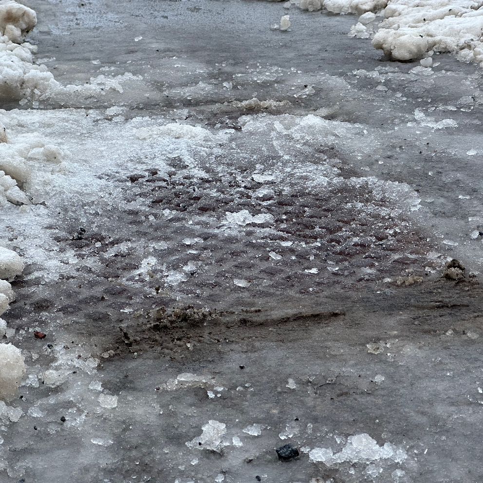 Vinterlandskap med smält is och snö på en väg, där en del av ett brunnslock är synligt under isen.