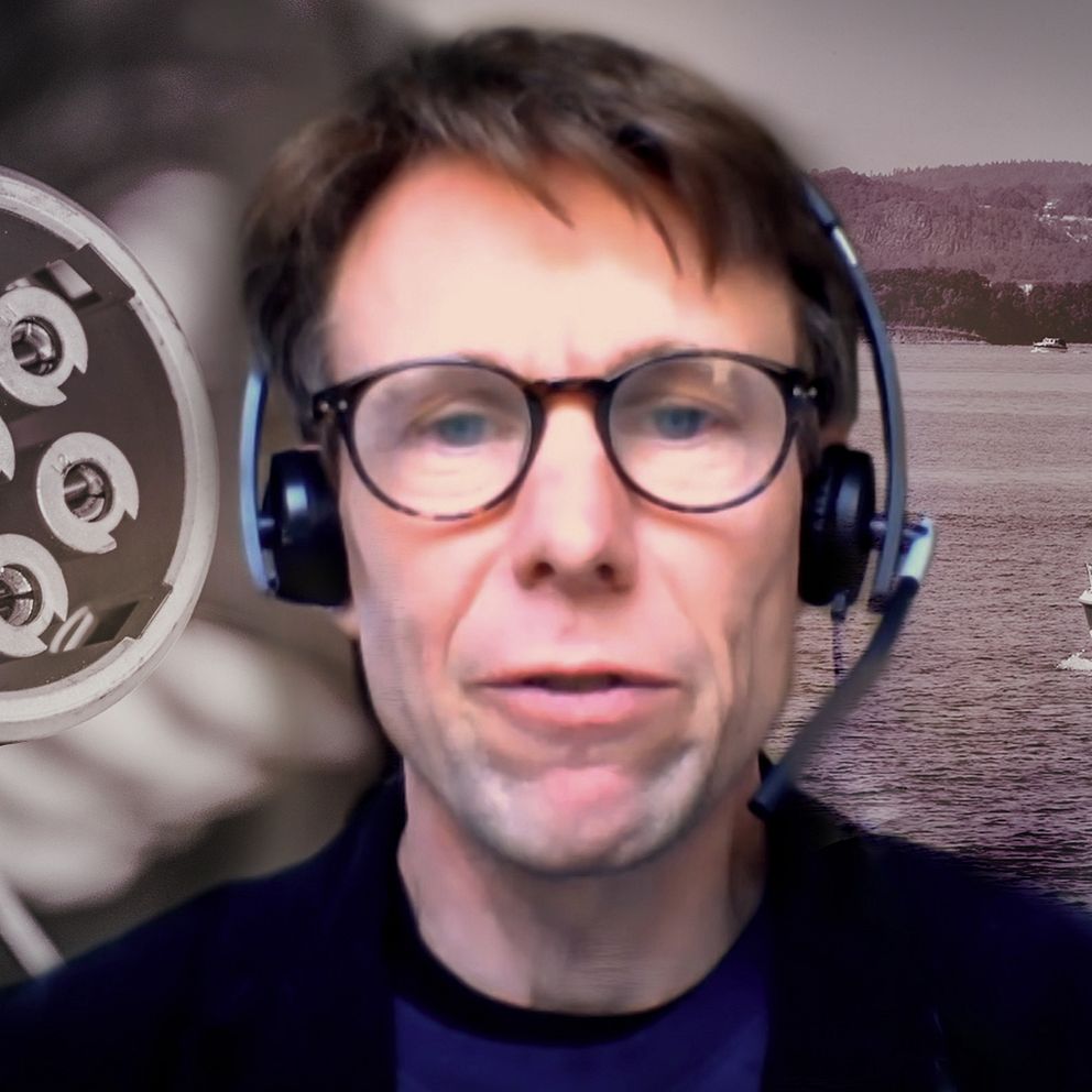 En person med hörlurar står framför en bakgrund där en båt syns som åker på vattnet, med en naturskön utsikt i bakgrunden. Till vänster i bilden syns en laddkontakt för elfordon.