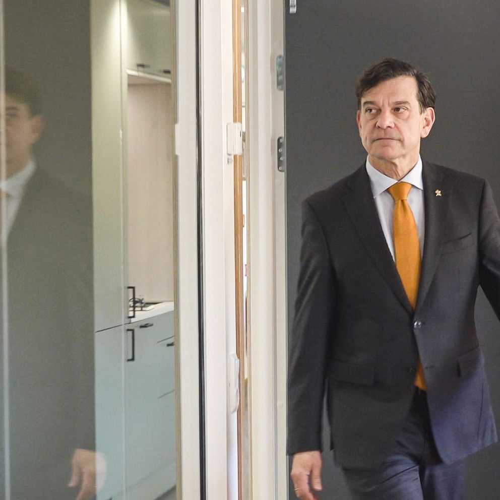 En man i kostym med orange slips går mot en dörr i en kontorsmiljö. Reflektionen av honom ses i det glasade elementet på vänster sida av bilden. Bakgrunden består av ljusa väggar och en dörr.
