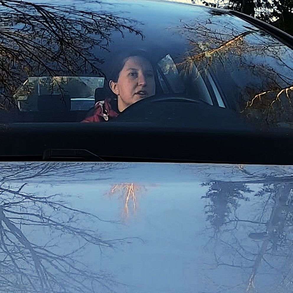 Träd speglar sig i vindruta och motorhuv, i bilen ser man Sofia Holmqvist