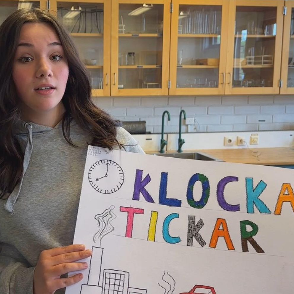 Amalia Lamnitchi står i ett klassrum på Gudmundråskolan i Kramfors. Hon har långt mörkt hår, grå huvtröja och håller i ett plakat med en klocka, industri och bil samt texten ”klockan tickar” i olika färger.
