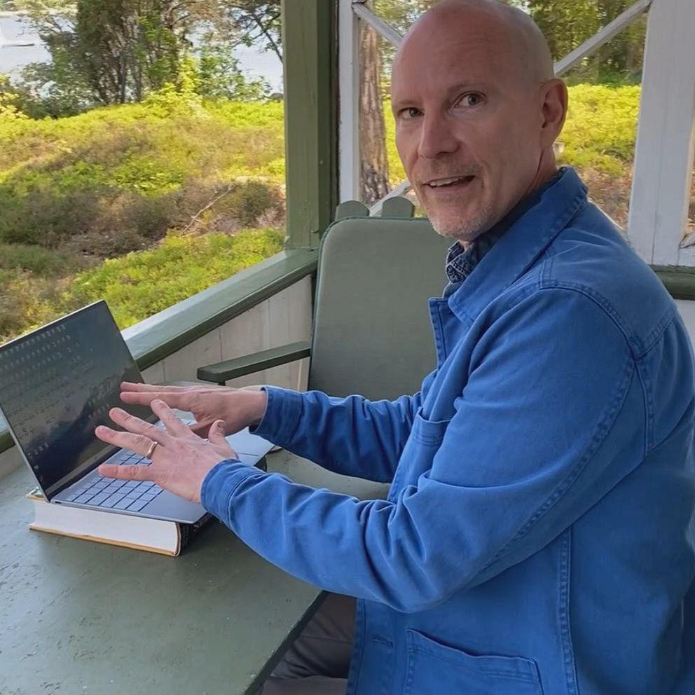 Man i blå skjorta sitter vid dator på veranda med utsikt över blåbärsris och hav med båtar. Peter Norrman, vd på Vulkan förlag, på Karön i Blekinge skärgård.