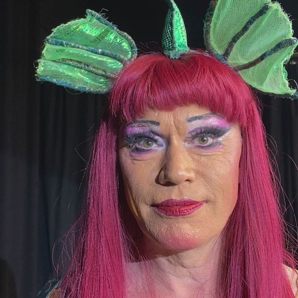 En dragshowartist ler och tittar in i kameran. Hen har gröna draköron, rött hår och glittrigt lila smink.
