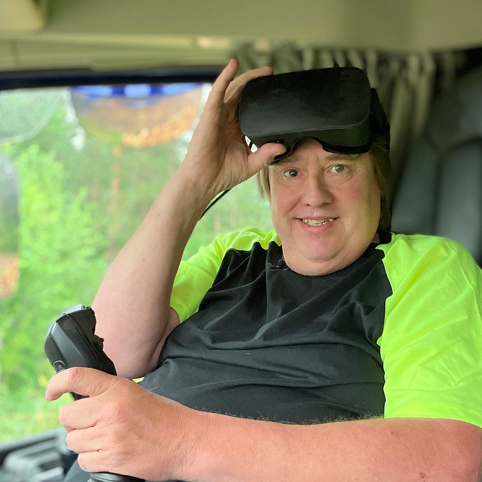 Thomas Bergander, man i svart och neongul t-shirt sitter i en lastbil med en joystick och ett par VR-glasögon i pannan.