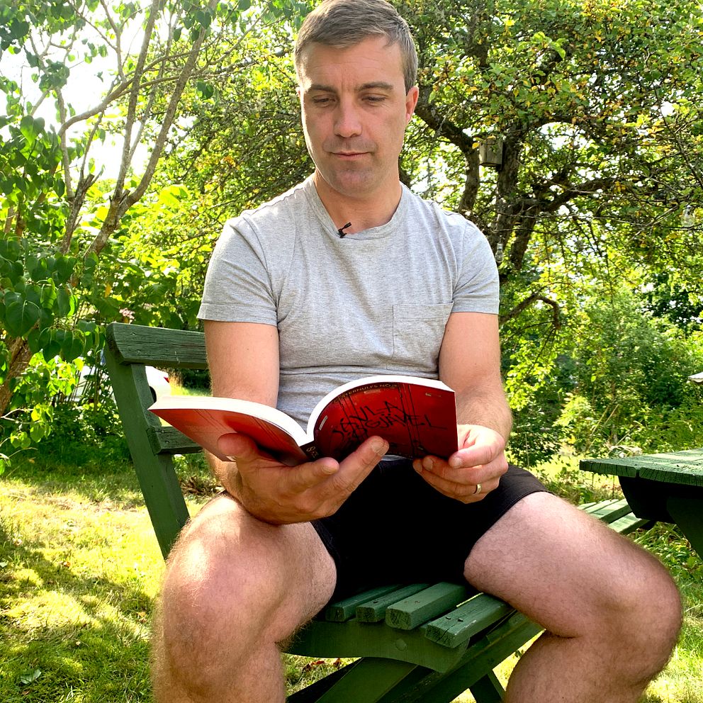 En man sitter på en stol i trädgård och läser en bok.