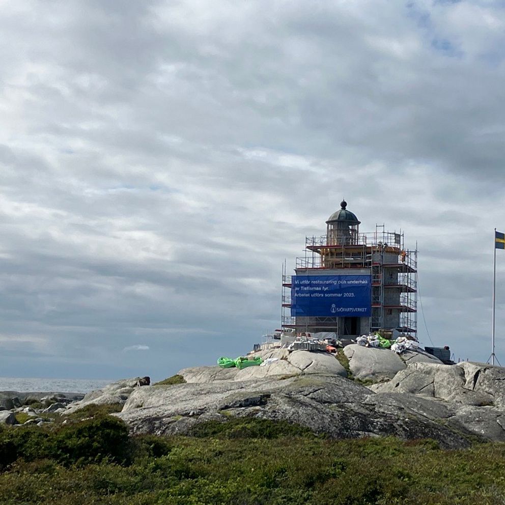 En fyr, Tistlarnas fyr, med byggnadsställning runtom står på en klippa. Bredvid fyren står en flaggstång med svenska flaggan hissad.