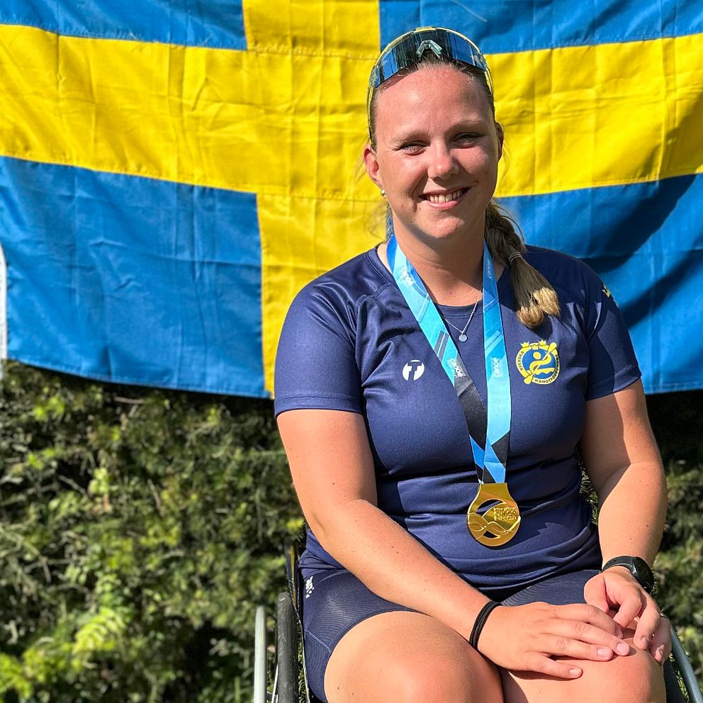 En parakanotist sitter i en rullstol framför svenska flaggan. Runt hennes hals hänger en guldmedalj.