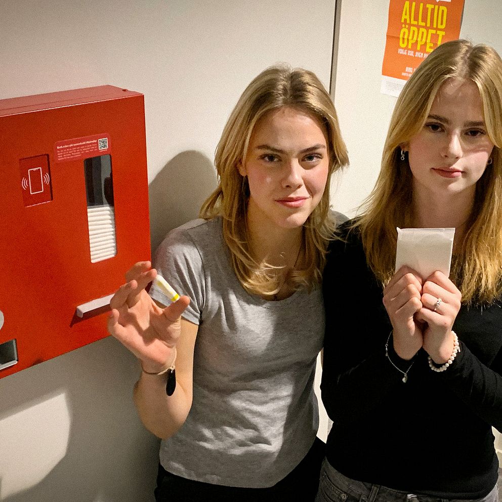 Eleverna Paulina och Nina håller upp mensskydd framför en röd automat.