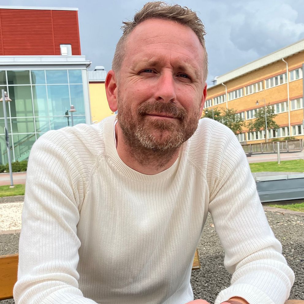 En man i vit tröja sitter på en bänk utomhus och tittar in mot kameran med ett litet leende.