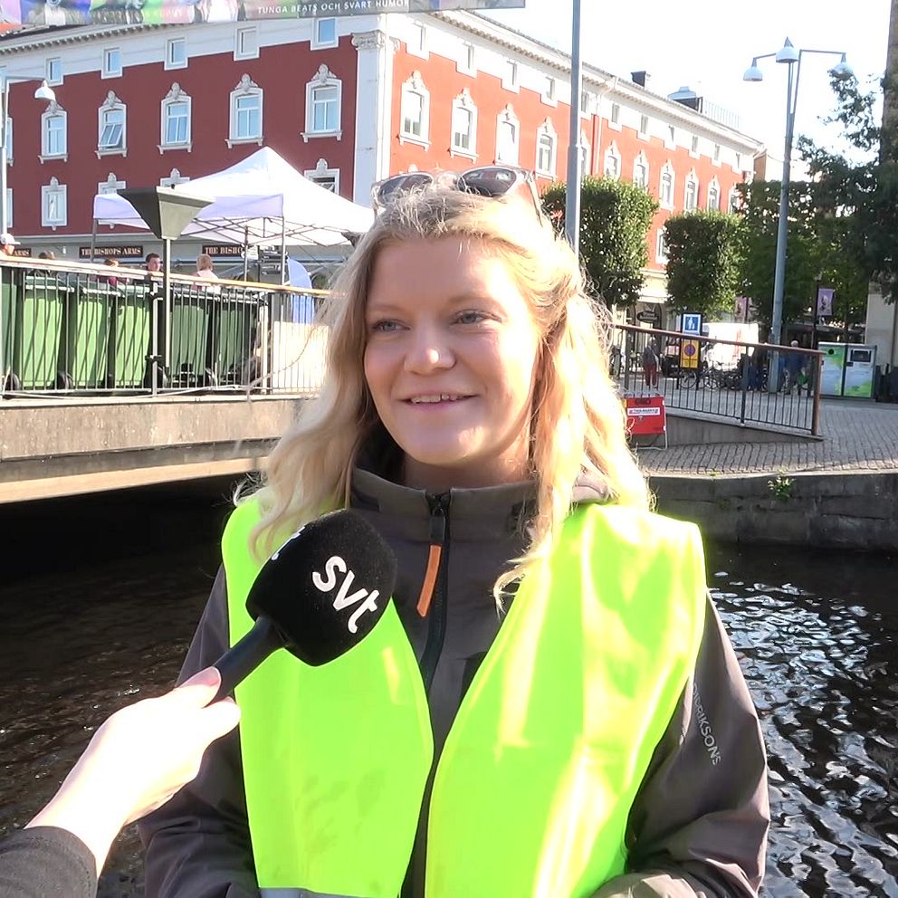 En kvinna med blont hår ler och tittar mot kameran. Hon står i centrala Jönköping och i bakgrunden syns kanalen i Munksjön och människor som går över vindbron.