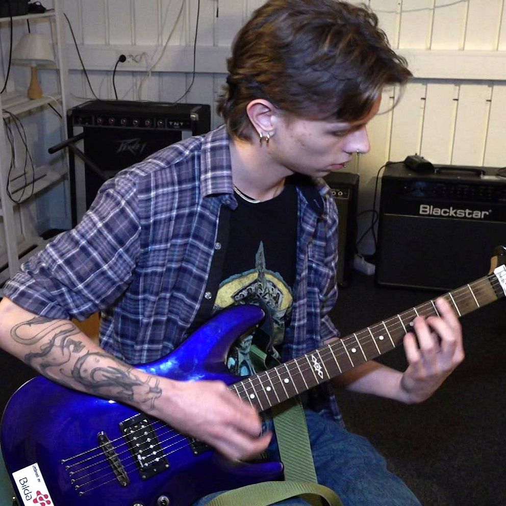 Ung man med mörkt hår och tatueringar spelar på en blå elgitarr.
