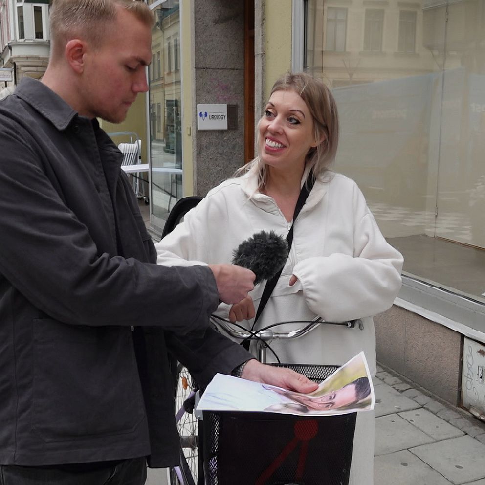 En man står i stadsmiljö iförd en grön skjortjacka. Han håller i en mikrofon och i en bild på en person som han visar upp för en blond kvinna i vit tröja som går med en cykel. Kvinnan skrattar lite lätt och tittar på mannen.