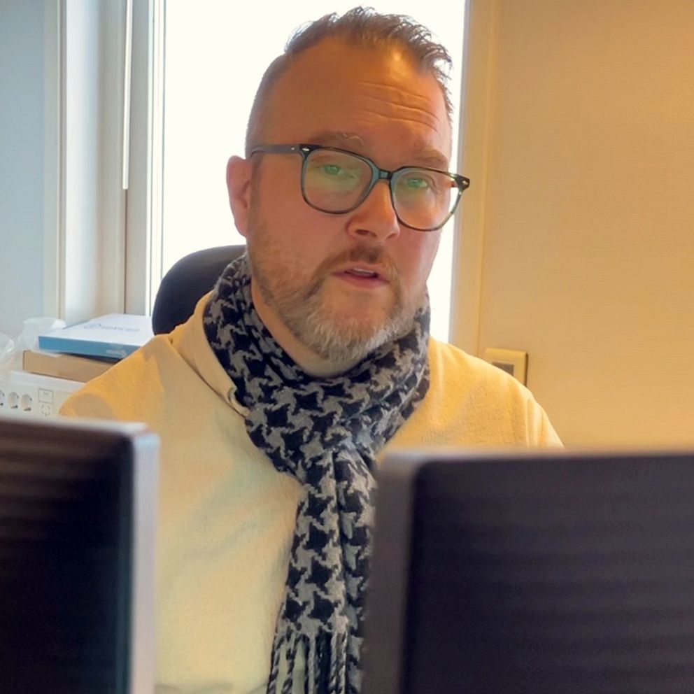 Stefan Rönnlund sitter vid ett skrivbord utrustat med två datorskärmar, med ett fönster i bakgrunden som släpper in naturligt ljus.