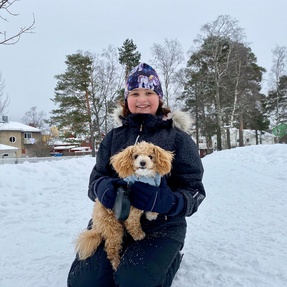Ett barn sitter i en snöig park och håller en lurvig liten hund. Träd och hus syns i bakgrunden under en mulen vinterdag.