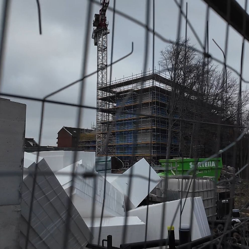 Byggarbetsplats med en kran och ett pågående byggprojekt omgiven av ett staket. I förgrunden syns staplade byggmaterial och stora vita isoleringsblock, medan en byggnad under uppförande reser sig i bakgrunden. Mörka moln skapar en dyster atmosfär.