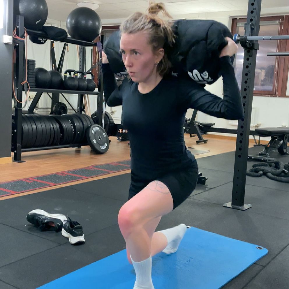 Kvinna utför en knäböj med vikt på axlarna i ett gymmiljö, med vikter och träningsutrustning i bakgrunden. Hon står på en blå träningsmatta och har på sig sportkläder.