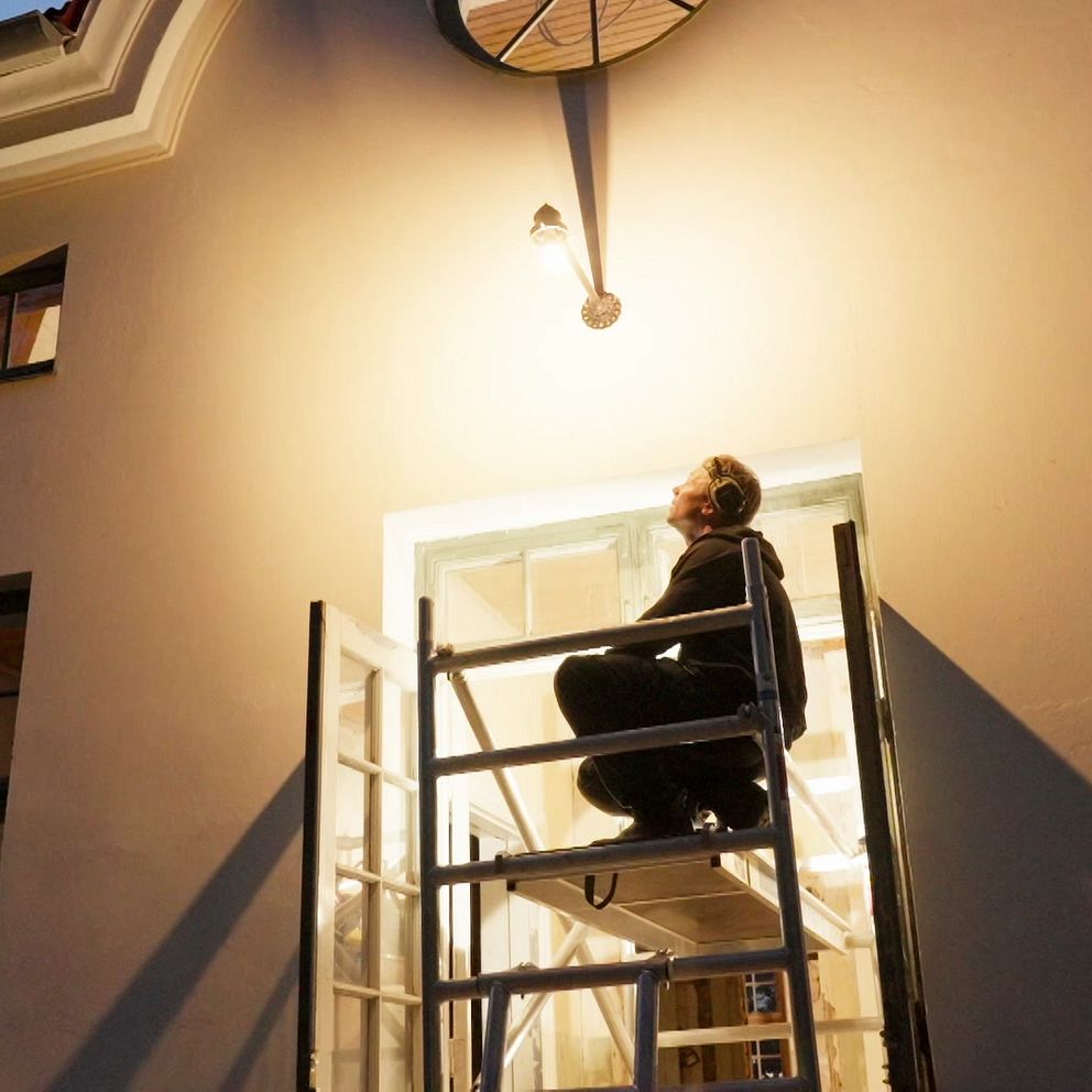 Person på byggställning monterar lampa över entré i skymning.