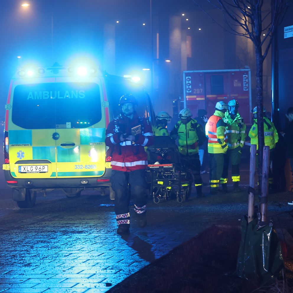 Ambulanspersonal och brandmän i reflexkläder står vid en olycksplats på en dimmig gata på natten. En ambulans med blåljus är synlig i bakgrunden.