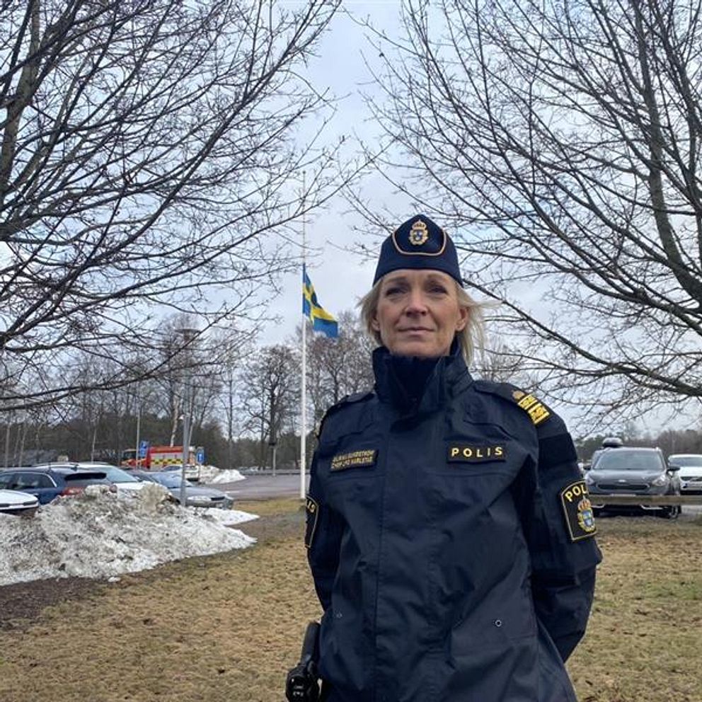 En polis i uniform står på en gräsyta med parkerade bilar i bakgrunden och en svensk flagga som vajar. Träd utan löv omger platsen, vilket antyder att det är vinter eller tidig vår. Snö kan ses samlad på marken.
