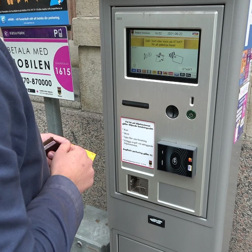 En person står vid en automat för betalning av parkering, där de håller en biljett eller kort i handen. Automaten har en digital skärm som visar olika options för betalning och instruktioner. I bakgrunden syns en informationsskylt om parkering.