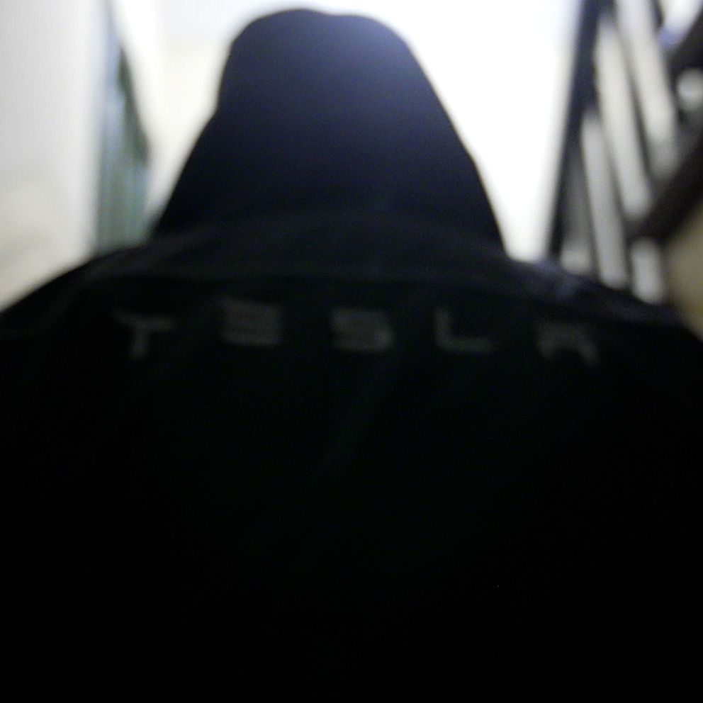 En person i en svart hoodie med texten ”TESLA” synlig bakifrån, står i en trappa med starkt ljus som kommer ovanifrån. Personen är anonym och står med ryggen mot kameran.