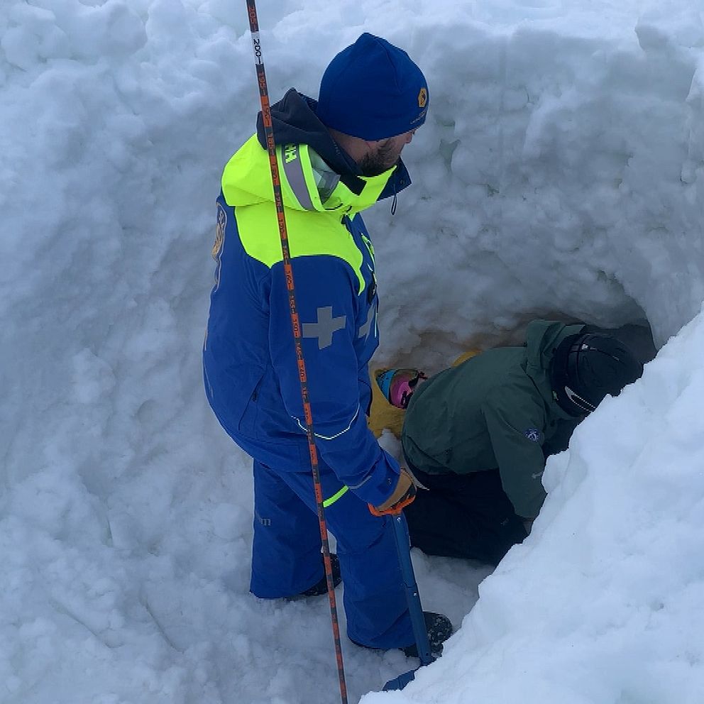 Två personer klädda i vinterkläder befinner sig i en grävd snögrop. En person står ovanför och håller i en stav medan den andra personen sitter i gropen. Omgivningen är täckt av snö.