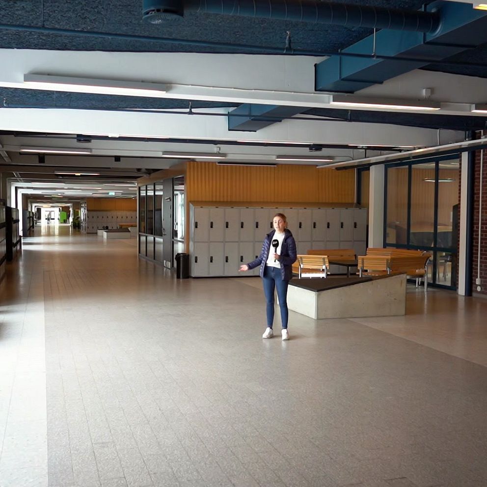 En kvinna står mitt i en lång, bred korridor med ljusa golv och högt i tak. Till höger finns en rad skåp och en dörr märkt ”WC”. Korridoren är tom förutom personen, och det finns bänkar längs fönstren till vänster.