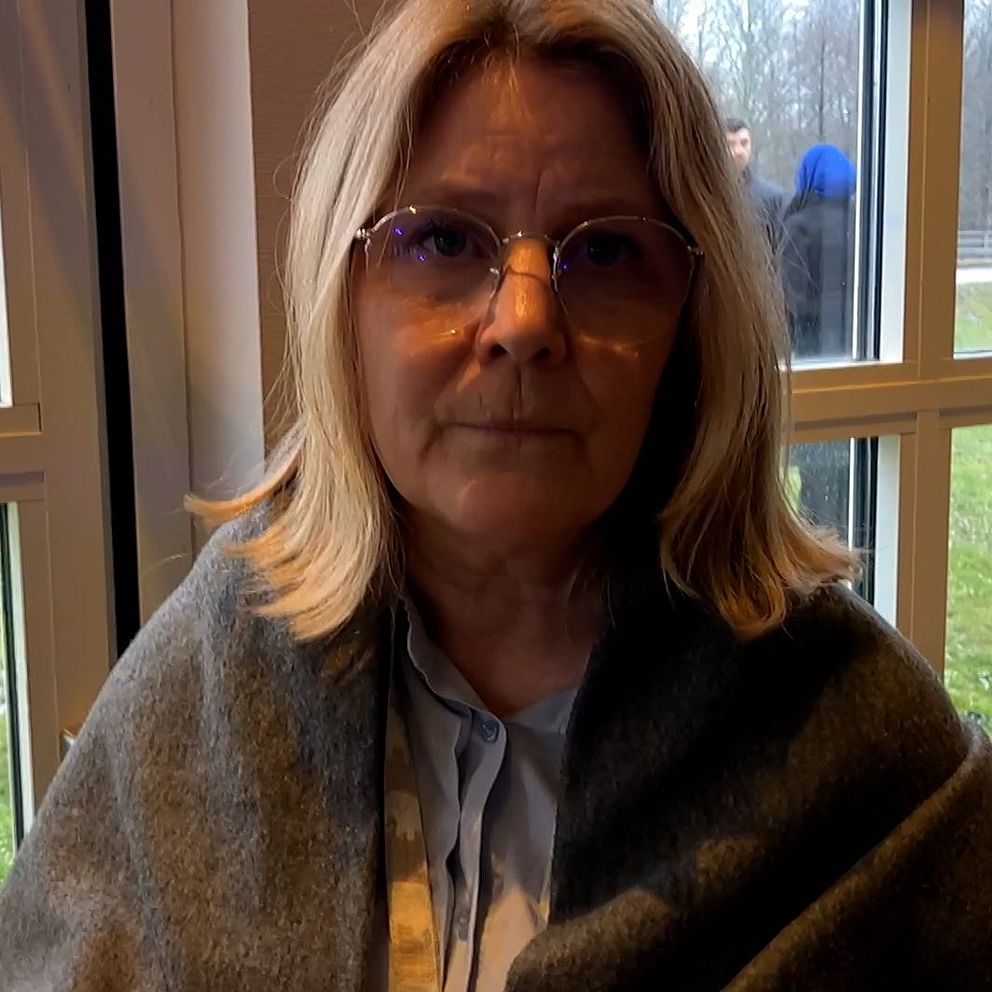 En person med blont hår sitter inomhus nära ett fönster och är insvept i en grå filt. I bakgrunden syns en skymt av en grön gräsmatta och människor som står utomhus.