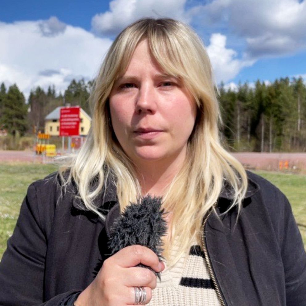 En person med mikrofon står utomhus med hus och skog i bakgrunden.