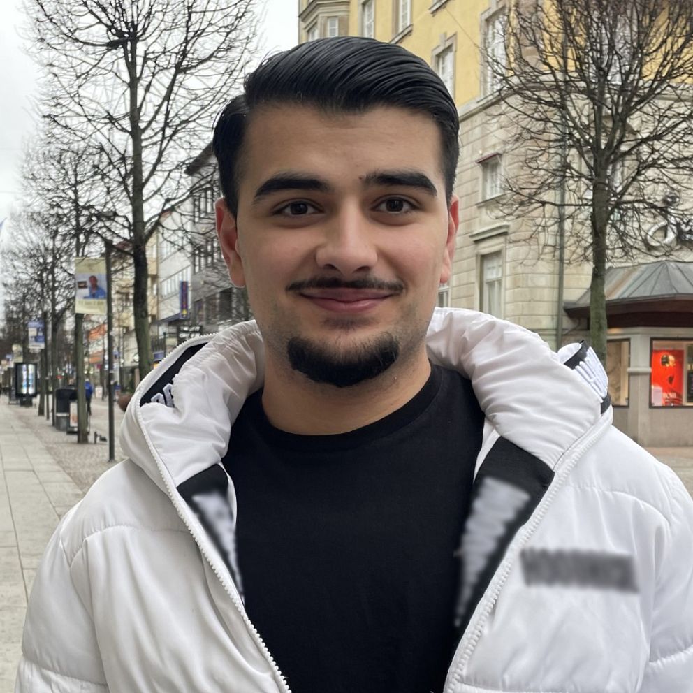 En man iförd en vit jacka står på en trottoar. Bakom syns en lång gata med butiker och restauranger.