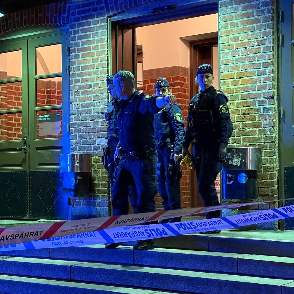 Poliser står vid en entré som är avspärrad med polisband. En manlig polis pekar mot kameran medan två andra poliser står bakom honom. Bilden är tagen på kvällen och har ett blåaktigt sken från belysningen.