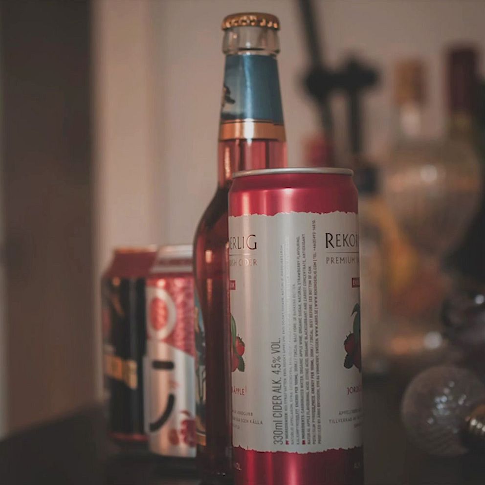 En uppsättning ciderburkar och flaskor på ett bord som inkluderar en röd burk med vit text i förgrunden, en flaska öl med en blårandig kapsyl bakom den, och en annan burk bakom den med ett mönster. I bakgrunden syns suddiga flaskor av olika slag.